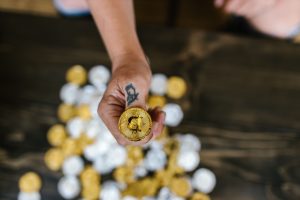 A person with a tattoo holding a gold Bitcoin amidst blurred coins signifying digital currency and wealth