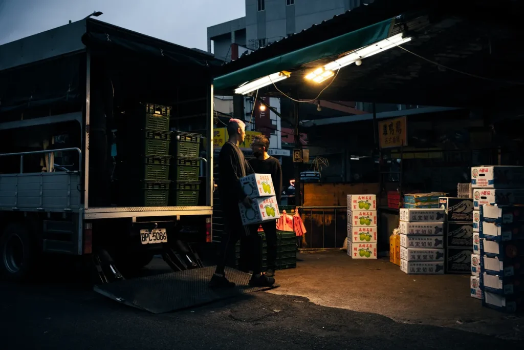 Workers unloading boxes from a delivery truck at an urban market during dusk