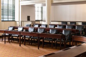 Empty courtroom seating with dark chairs, wooden rails and tall windows