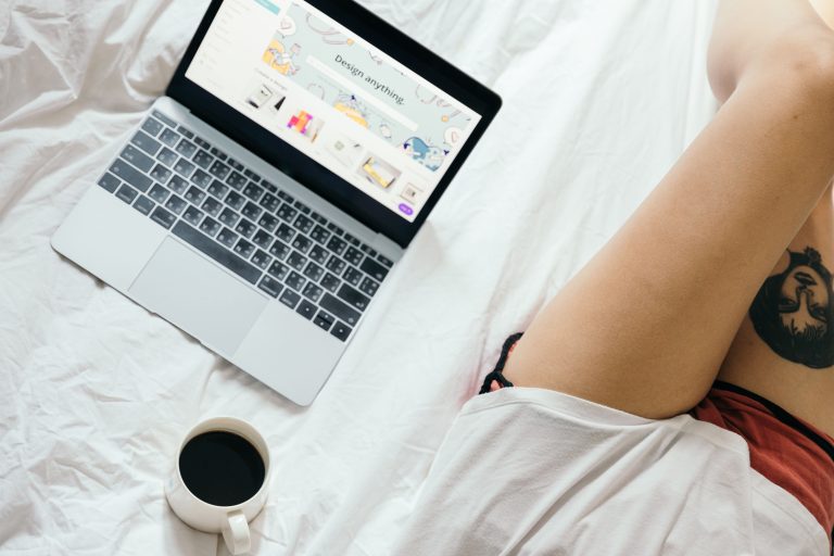 From above of unrecognizable woman with tattoo on leg browsing netbook and drinking coffee while resting on bed in cozy bedro