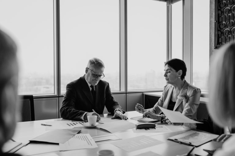 Two people review papers at a meeting table beside tall windows