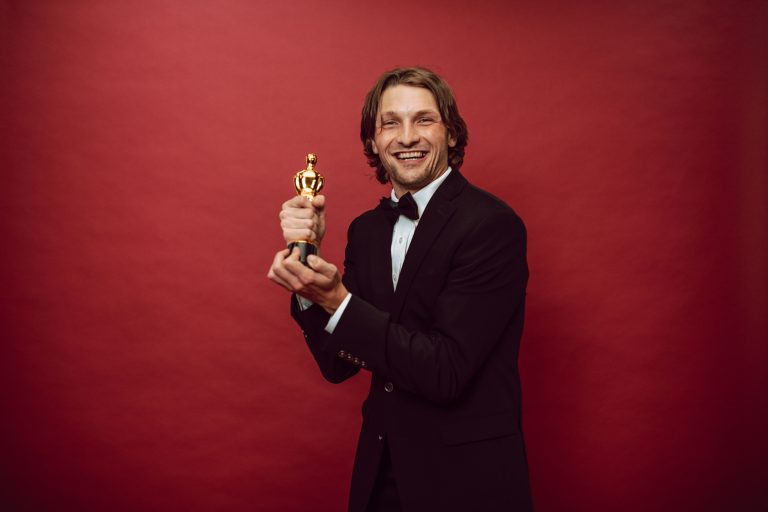 Smiling man in a tuxedo holding a trophy against a red background, celebrating success